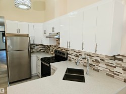 Galley kitchen featuring white cabinetry, light-toned countertops, a stainless steel double basin sink with a high-arch faucet, and a rectangular tile backsplash - 