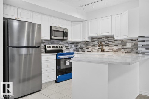 White cabinetry with panel detailing, stainless steel appliances, mosaic tile backsplash, light-toned countertop, and track lighting - #1102 10909 103 Av Nw, Edmonton, AB - Indoor Photo Showing Kitchen