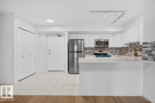 White cabinetry with gold-tone hardware, stainless steel appliances, a tiled backsplash, and a breakfast bar with a solid surface countertop - #1102 10909 103 Av Nw, Edmonton, AB - Indoor Photo Showing Kitchen With Upgraded Kitchen