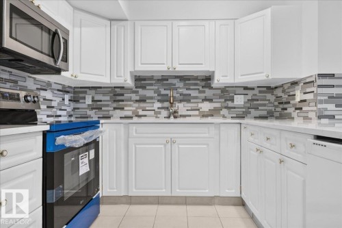 White cabinetry with gold-toned hardware, stainless steel appliances, light-toned countertops, and a gray tile backsplash - #1102 10909 103 Av Nw, Edmonton, AB - Indoor Photo Showing Kitchen
