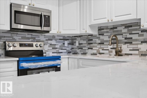 White cabinetry with gold-tone hardware, stainless steel microwave, stainless steel range, mosaic tile backsplash, and light-toned countertop - #1102 10909 103 Av Nw, Edmonton, AB - Indoor Photo Showing Kitchen