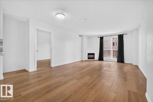 Wood-finish flooring, white painted walls, electric fireplace with white mantel, ceiling-mounted light fixture, and large window - #1102 10909 103 Av Nw, Edmonton, AB - Indoor Photo Showing Living Room With Fireplace