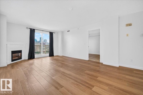 Expansive living area featuring wood-finish flooring, a white mantel fireplace, and a large window with city architecture visible - #1102 10909 103 Av Nw, Edmonton, AB - Indoor Photo Showing Living Room With Fireplace