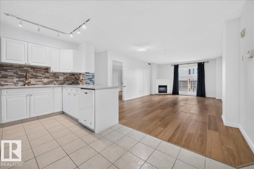 White cabinetry with gold-tone hardware, a mosaic tile backsplash, a built-in dishwasher, a white fireplace, and wood-finish flooring - #1102 10909 103 Av Nw, Edmonton, AB - Indoor Photo Showing Kitchen