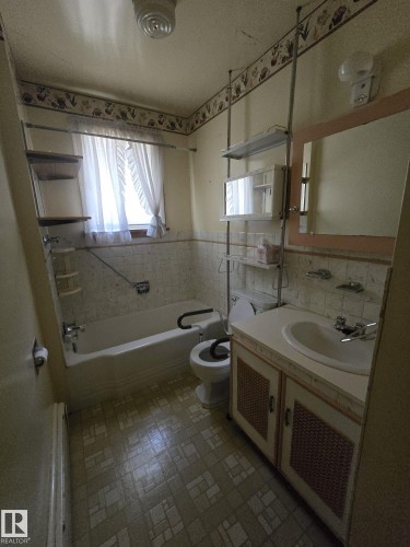Bathroom featuring a window with sheer curtains, a built-in bathtub, a toilet with an attached support bar, a vanity with an integrated sink, and patterned floor tile - 9839 81 Avenue Nw, Edmonton, AB - Indoor Photo Showing Bathroom
