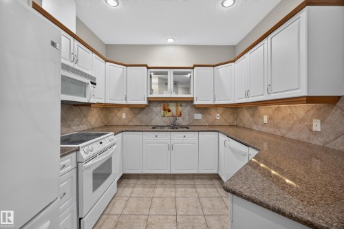 Kitchen featuring white cabinetry with wood trim, speckled countertops, tile backsplash, and tile flooring - #401 10178 117 St Nw, Edmonton, AB - Indoor Photo Showing Kitchen With Double Sink