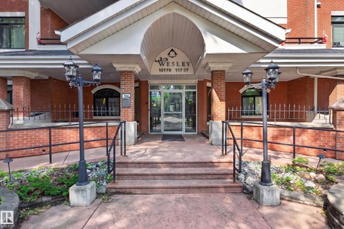 Brick exterior with a covered entry, featuring a multi-pane glass door, paved steps, an accessible ramp with a handrail, and twin decorative street lamps - #401 10178 117 St Nw, Edmonton, AB - Outdoor