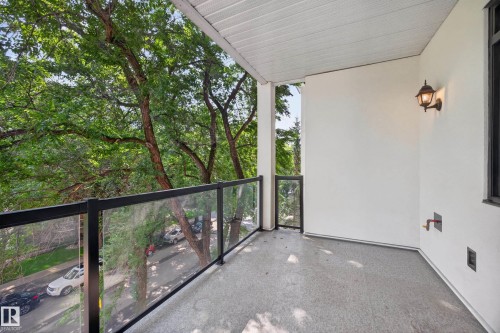 Outdoor balcony featuring a glass and metal railing, textured flooring, white stucco walls, a classic wall sconce, and a visible spigot - #401 10178 117 St Nw, Edmonton, AB - Outdoor With Exterior