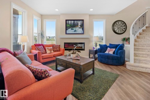 Living area with warm neutral walls, recessed lighting, wood-finish flooring, a contemporary electric fireplace, and a curved staircase with white spindles - 7135 Edgemont Way Nw, Edmonton, AB 