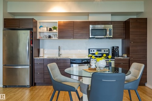 Contemporary kitchen featuring wood-finish cabinetry, stainless steel appliances, white countertops, and a glass-top dining table - #407 9523 160 Av Nw, Edmonton, AB - Indoor