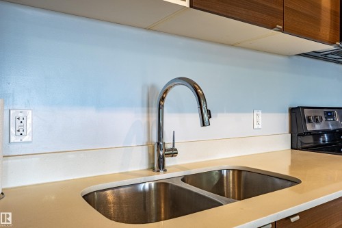 Kitchen featuring a dual basin stainless steel sink with a high-arch chrome faucet, solid surface countertops, and modern wood-finish cabinetry - #407 9523 160 Av Nw, Edmonton, AB - Indoor Photo Showing Kitchen With Double Sink