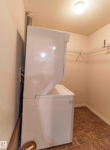 This dedicated laundry area features a white stacked washer and dryer, tile flooring, and wire shelving for storage, all illuminated by a ceiling-mounted light fixture - #1111 7339 South Terwillegar Dr Nw, Edmonton, AB - Indoor Photo Showing Laundry Room