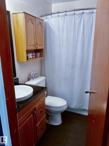 Bathroom featuring a wood-finish vanity with a vessel sink, dark countertop, toilet, and a bathtub with a shower curtain - 11739 129 Street Nw, Edmonton, AB 