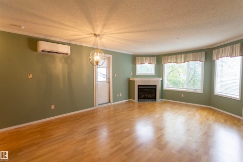 Hardwood-finish flooring, built-in fireplace with a light-toned mantel, multiple windows, wall-mounted air conditioning unit, and a decorative ceiling light fixture - #204 2420 108 St Nw, Edmonton, AB - Indoor Photo Showing Living Room With Fireplace