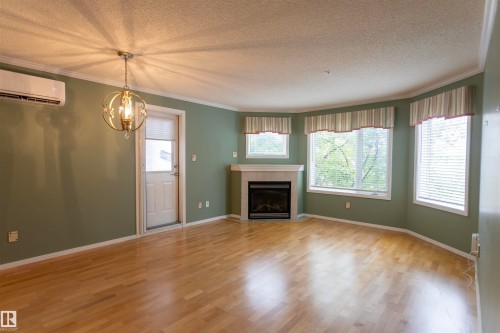 Hardwood-finish flooring, corner fireplace with tile surround, curved wall with multiple windows, crown molding, and a built-in air conditioning unit - #204 2420 108 St Nw, Edmonton, AB - Indoor Photo Showing Living Room With Fireplace