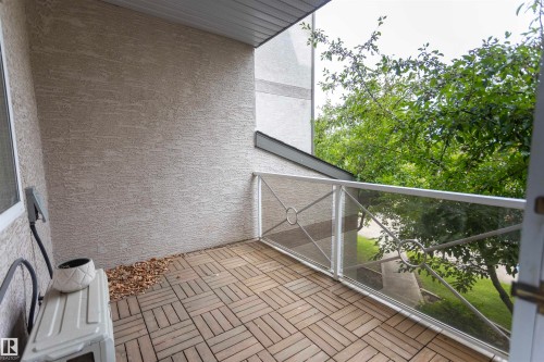 Balcony featuring wood-finish floor tiles, stucco exterior, and a glass-paneled railing with decorative metalwork - #204 2420 108 St Nw, Edmonton, AB - Outdoor With Exterior