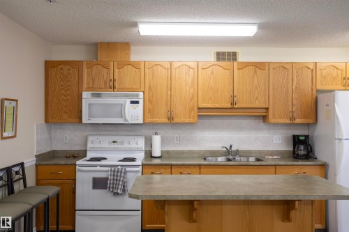 Kitchen featuring wood cabinetry, white appliances, a tile backsplash, a kitchen island, and a dual basin stainless steel sink - #204 2420 108 St Nw, Edmonton, AB - Indoor Photo Showing Kitchen With Double Sink