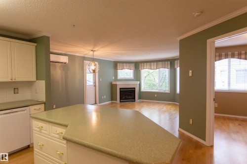 Kitchen island with integrated cabinetry and light-toned countertop, wood-finish flooring, and a fireplace with a mantel - #204 2420 108 St Nw, Edmonton, AB - Indoor Photo Showing Kitchen With Fireplace