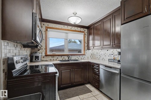 Dark wood-finish cabinetry, stainless steel appliances, light-toned countertop, mosaic tile backsplash, and a double basin sink - 4323 114B Street Nw, Edmonton, AB - Indoor Photo Showing Kitchen With Double Sink