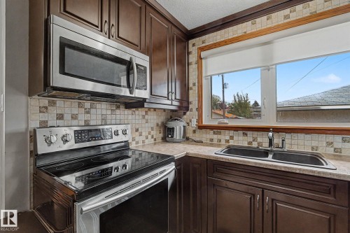 Kitchen featuring dark wood-finish cabinetry, stainless steel appliances, a tiled backsplash, and a double basin sink with a window above - 4323 114B Street Nw, Edmonton, AB - Indoor Photo Showing Kitchen With Double Sink