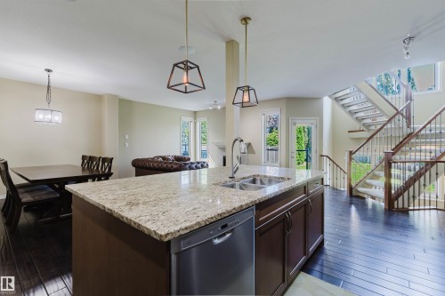 Kitchen island with integrated sink and dishwasher, wood-finish flooring, two-story staircase with wrought iron balusters, and transom windows - 864 Armitage Wynd Sw, Edmonton, AB - Indoor Photo Showing Kitchen With Double Sink