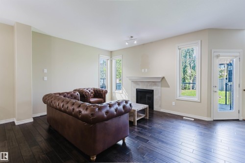 Spacious living area featuring dark wood-finish flooring, a stone fireplace with mantel, white trim, multiple windows, and a glass-paneled exterior door - 864 Armitage Wynd Sw, Edmonton, AB - Indoor Photo Showing Living Room With Fireplace