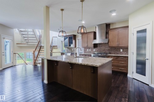 Kitchen island with a granite countertop, dark wood-finish flooring, stainless steel range hood, pendant lighting, and a glass-paneled door - 864 Armitage Wynd Sw, Edmonton, AB - Indoor Photo Showing Kitchen With Upgraded Kitchen
