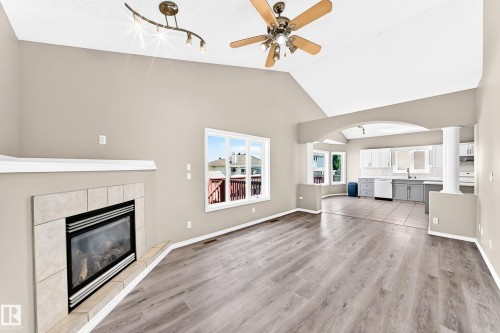 Vaulted ceilings with a ceiling fan, modern fireplace with tile surround, wood-finish flooring, and an arched entry to a kitchen with white cabinetry - 16227 64 Street Nw, Edmonton, AB - Indoor Photo Showing Living Room With Fireplace