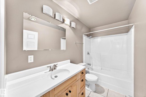 Bathroom featuring a wood-finish vanity with a white countertop and integrated sink, a large wall-mounted mirror, a tub/shower combination with a white surround, and light-toned tile flooring - 16227 64 Street Nw, Edmonton, AB - Indoor Photo Showing Bathroom