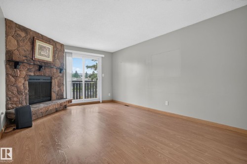 Living area featuring a stone-clad fireplace with a matching hearth, wood-finish flooring, light grey wall paint, and a sliding glass door opening to a balcony with metal railings - 10644 158 Avenue Nw, Edmonton, AB - Indoor Photo Showing Living Room With Fireplace