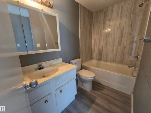 White vanity with integrated sink, three-panel mirrored medicine cabinet, white bathtub with tiled surround, and wood-finish flooring - 10829 167A Avenue Nw, Edmonton, AB - Indoor Photo Showing Bathroom