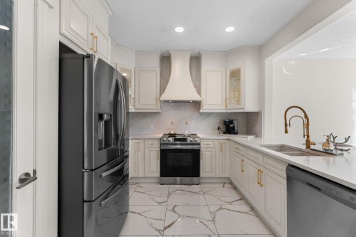 U-shaped kitchen featuring white cabinetry with gold-finish hardware, stainless steel appliances, a range hood, and large format floor tiling - 5415 167A Avenue Nw, Edmonton, AB - Indoor Photo Showing Kitchen With Double Sink