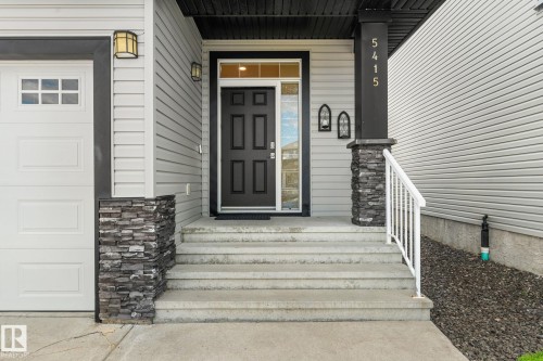 Front exterior featuring light gray siding, a black paneled entry door with sidelights, a white garage door with upper window inserts, concrete steps with a white metal handrail, and stone accent pillars - 5415 167A Avenue Nw, Edmonton, AB - Outdoor