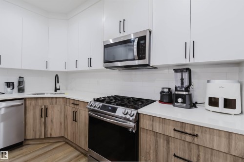 Kitchen featuring white upper cabinetry, wood-finish lower cabinetry, a stainless steel gas range, a stainless steel microwave, and white countertops - 104 Meadowview Way, Leduc, AB - Indoor Photo Showing Kitchen With Upgraded Kitchen