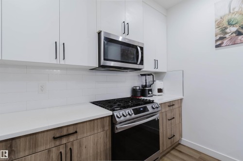 White upper cabinetry with black hardware, wood-finish lower cabinetry, white subway tile backsplash, white countertops, and stainless steel microwave and range - 104 Meadowview Way, Leduc, AB - Indoor Photo Showing Kitchen With Upgraded Kitchen