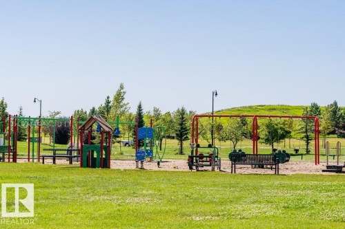 Community playground featuring multiple play structures, swings, and benches, set against a backdrop of green space and mature trees - 1539 Kinross Road Nw, Edmonton, AB - Outdoor