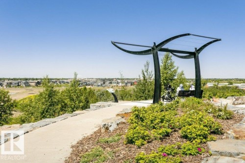 Paved pathway with stone retaining walls, leading to a modern shaded seating area, surrounded by landscaped plantings and distant residential structures - 1539 Kinross Road Nw, Edmonton, AB - Outdoor With View