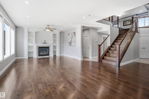 Wood-finish flooring throughout, featuring a fireplace with built-in shelving, a ceiling fan, and a grand staircase with wood railing - 427 Windermere Road Nw, Edmonton, AB - Indoor Photo Showing Living Room With Fireplace