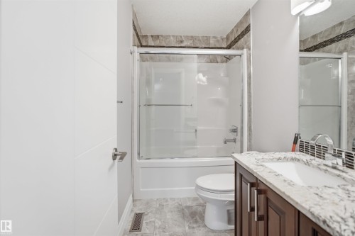 White panel door with satin nickel hardware, a shower-over-tub unit with sliding glass doors, a vanity with a white composite sink and chrome faucet, a stone-look countertop, and dark wood-finish cabinetry - 427 Windermere Road Nw, Edmonton, AB - Indoor Photo Showing Bathroom