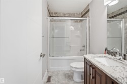 White panel door with satin nickel hardware, a shower-over-tub unit with sliding glass doors, a vanity with a white composite sink and chrome faucet, a stone-look countertop, and dark wood-finish cabinetry - 