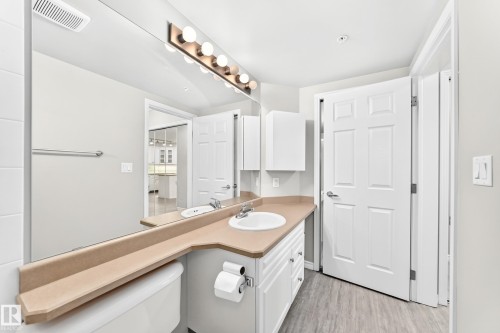 Bathroom featuring a full-width vanity mirror, a single basin sink, white cabinetry, a wall-mounted light fixture, and wood-finish flooring - #137 9704 174 St Nw, Edmonton, AB - Indoor Photo Showing Bathroom