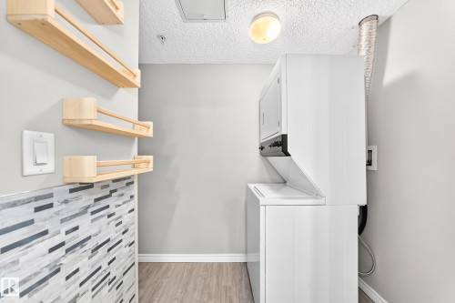 Stacked laundry appliances, wood-finish flooring, light grey wall paint, and decorative wall shelving - #137 9704 174 St Nw, Edmonton, AB - Indoor Photo Showing Laundry Room
