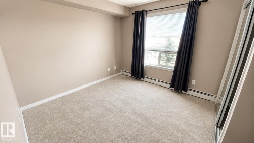 Neutral-toned carpeting, window with blinds and dark drapery, baseboard heating, and a mirrored closet door - #301 13907 136 St Nw, Edmonton, AB - Indoor Photo Showing Other Room