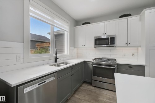 Kitchen featuring two-tone cabinetry, stainless steel appliances, a double basin sink, solid surface countertops, and wood-finish flooring - 7603 Summerside Grande Boulevard Sw, Edmonton, AB - Indoor Photo Showing Kitchen With Double Sink