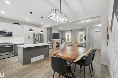 Open-concept living space with wood-finish flooring, white cabinetry, stainless steel appliances, and a stone-clad fireplace - 7603 Summerside Grande Boulevard Sw, Edmonton, AB - Indoor