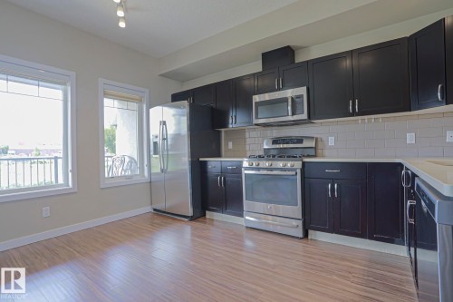 Contemporary kitchen featuring dark cabinetry, stainless steel appliances, light countertops, subway tile backsplash, and wood-finish flooring - #13 5873 Mullen Pl Nw, Edmonton, AB - Indoor Photo Showing Kitchen