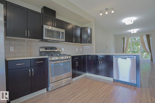 Kitchen featuring dark wood cabinetry, stainless steel appliances, light-toned countertops, subway tile backsplash, and wood-finish flooring - #13 5873 Mullen Pl Nw, Edmonton, AB - Indoor Photo Showing Kitchen