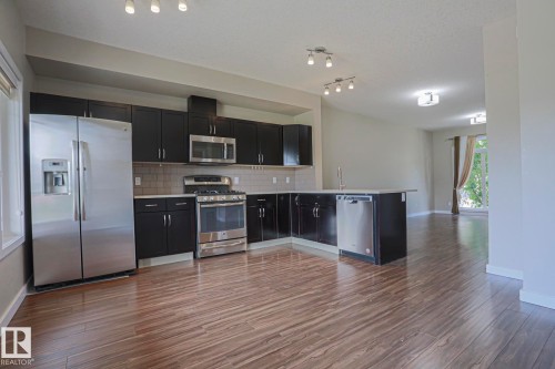 Kitchen featuring dark cabinetry, stainless steel appliances, an island with an undermount sink, a tiled backsplash, and wood-finish flooring - #13 5873 Mullen Pl Nw, Edmonton, AB - Indoor Photo Showing Kitchen