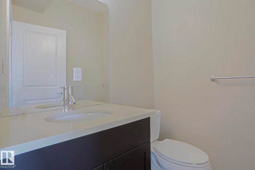 White solid surface countertop, integrated white sink, chrome faucet, dark wood-finish vanity, and wall-mounted mirror - #13 5873 Mullen Pl Nw, Edmonton, AB - Indoor Photo Showing Bathroom