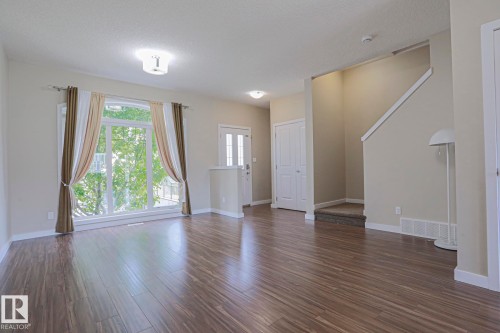 Spacious living area featuring wood-finish flooring, a large picture window, and neutral wall tones - #13 5873 Mullen Pl Nw, Edmonton, AB - Indoor